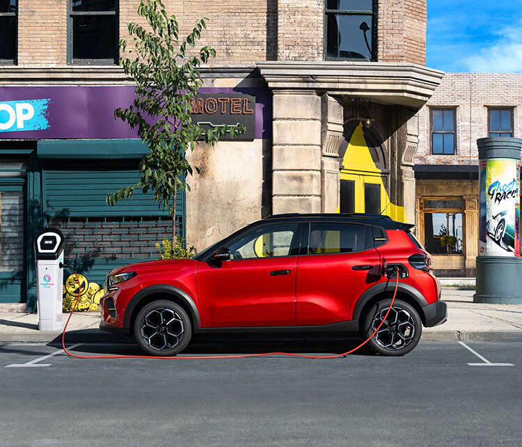 Новий Citroën ë-C3 червоного кольору заряджається на громадській зарядній станції, вигляд з лівого боку